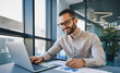 © Pixelowy_czarodziej - A smiling young man in a suit sits in the office at a desk and works on a laptop, typing on a keyboard, texting, working with data