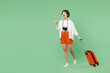 © ViDi Studio - Full body traveler woman wears casual clothes hold suitcase bag walk go point index finger aside isolated on plain green background. Tourist travel abroad in free time rest. Air flight trip concept.