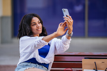 young woman in casual attire is enjoying her day, posing and taking selfies while sitting on a bench outdoors. Shopping bags and a bright, cheerful atmosphere surround her