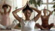 © artitwpd - A young woman sits in a yoga pose with her eyes closed and her hands together at the top of her head. She is wearing a white t-shirt and black leggings. There are two other women in the background.