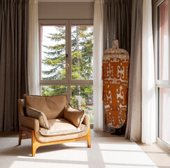 Detail of a modern living room with large windows that let in a lot of light and look out over large plants outside. There is a designer armchair with cushions on it
