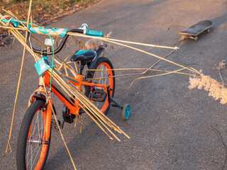 Two-wheeled orange and blue child bike with extra wheels carrying bulrush and pulling a skate board on a rope behind. Child playing active creative games outdoors in a park.