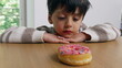 © Marco - A young boy sitting at a table, hands clasped, intently gazing at a colorful sprinkled donut, demonstrating delayed gratification with a thoughtful and curious expression
