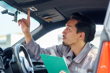 Wall Mural - Asian male insurance officer in uniform using clipboard to inspect car interior. Pointing towards car roof light, focusing on thorough inspection during claim process. Represents attention to detail,