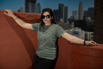 A stylish person in sunglasses, green shirt, and black shorts leans against a red rooftop wall with an urban cityscape in the background, exuding a casual and urban vibe.