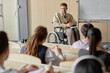 © Seventyfour - Adult male professor using wheelchair lecturing university students while holding microphone speaking with audience