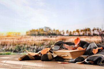 Poster - Autumn composition of black blanket and book and  autumn tree leaves on wooden table with copy space for products. Autumn season blurred forest landscape background.