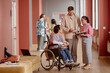 © Seventyfour - Adult male professor answering question in friendly informal conversation with male student using wheelchair and smiling female learner in pink university hallway