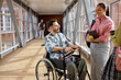 © Seventyfour - Side view of smiling Middle Eastern male student using wheelchair friendly talking to female classmates in university hallway with large windows, copy space