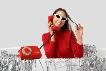 Poster - Happy young fashionable woman with retro phone and silver tinsel sitting in bathtub on grey background