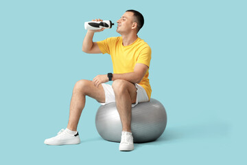 Wall Mural - Sporty young man drinking water on fitball against blue background