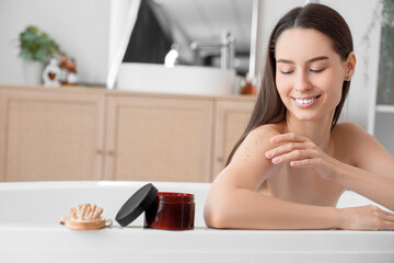 Canvas Print - Beautiful young happy woman with jar of body scrub and massage brush sitting in bathroom