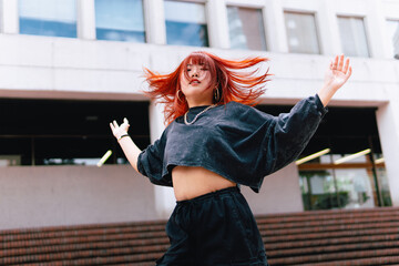 Wall Mural - An Asian woman with vibrant red hair dances dynamically in front of a modern urban building