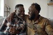© vadosloginov - Two Black men are sitting on a couch together, both laughing and looking at each other. They are indoors, in a home or apartment. The man on the right has his arm around the man on the left, and they