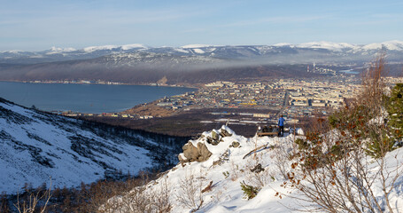 Wall Mural - View from the mountain to the city of Magadan. Tourists admire the city from the snow-covered slope of the hill. Young men sit on a bench and look into the distance. Magadan region, Far East of Russia