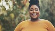 © red_orange_stock - A plus-sized woman smiles confidently outdoors, surrounded by lush greenery under bright sunlight