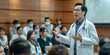 © Aigo labs - A doctor in a white coat and stethoscope is leading a medical conference, presenting information to a group of people wearing face masks.. asian man
