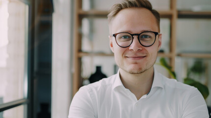 Wall Mural - A cheerful young man with glasses and a white shirt smiles confidently in a sunny room with bookshelves behind him.