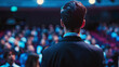 © mahamad - Rear view of people in the audience at a conference hall, with a speaker giving a talk at a business event.