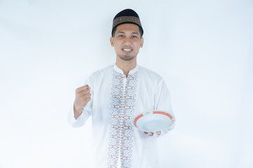 Cheerful young Asian Muslim man in cap doing a successful win gesture with clenched fist while presenting an empty plate and copy space over it