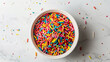 © thecreativesupplies - An overhead view of a bowl containing rainbow jimmies, centered on a white surface