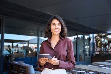 Portrait of middle eastern businesswoman using tablet pc app for work standing at office business building outdoors. Smiling indian or latin hispanic woman holding digital computer, looking at camera