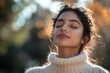 © Harvinder - A young beautiful Indian woman is standing outside in a park in a white sweater and with her eyes closed is taking deep breaths and enjoying herself. Close-up, Generative AI