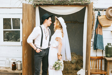 Wall Mural - wedding boho style ceremony young couple newlyweds bride in white dress with veil and groom in hat and jeans with suspenders hugging and having fun at barn of country house, shot in motion