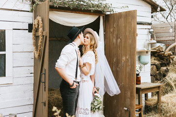 wedding boho style ceremony young couple newlyweds bride in white dress with veil and groom in hat and jeans with suspenders hugging and having fun at barn of country house, shot in motion