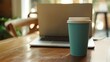 © Galib - A blue takeaway coffee cup on a wooden table with a laptop computer behind it.