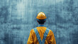 © Nima - Construction Worker Facing Concrete Wall, Back View - A construction worker in a yellow jumpsuit and hard hat stands facing a concrete wall, symbolizing safety, construction, industry, hard work