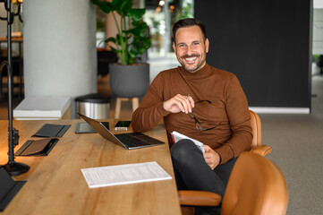 Wall Mural - Portrait of smiling male professional holding eyeglasses and working over laptop at desk in office