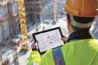 © JR-50 - Construction worker wearing a hard hat and safety vest, looking at a tablet computer on a construction site.