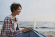 © ViDi Studio - Side view young IT woman she wear shirt casual clothes hold use work on laptop pc computer chatting sits rest on sea ocean sand shore beach outdoor seaside in summer day free time. Lifestyle concept.