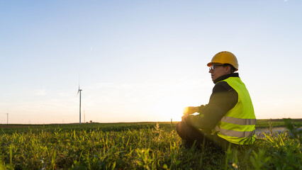 Canvas Print - Worker sits on the grass and looks at wind turbines