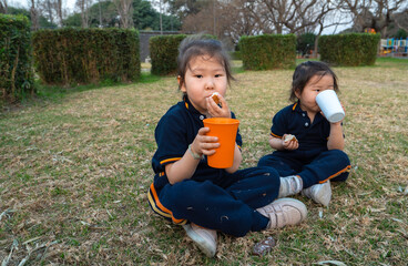 happy little girls sit, smile and eat on the grass in the park