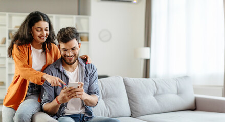 Happy young indian couple using smartphone, wife pointing at cellphone screen, sitting on sofa in living room interior at home. New app ad, banner