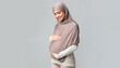 © Prostock-studio - Enjoying pregnancy. Pregnant muslim woman in headscarf tenderly touching her belly, embracing tummy over gray background with empty space, panorama