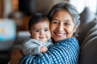 © Carmen - Senior Hispanic grandmother holding her grandchild, while sitting on cozy sofa, copy space