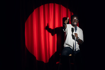 Wall Mural - Happy young african american man comic tells stand up monologue on stage with red curtains. Concept humor standup comedian show open mike