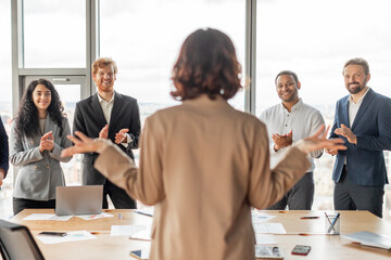 Aufkleber - A woman stands in front of a group of colleagues who are all clapping for her in a modern office setting. She appears to be accepting their praise and appreciation, smiling as she looks at them.