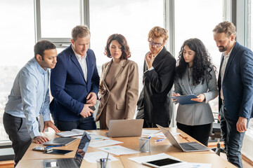 Aufkleber - A group of diverse business professionals in formal attire gather around a table in a modern office space. They are engaged in a collaborative meeting, discussing documents and a laptop