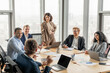 © Prostock-studio - A group of diverse business professionals are seated around a large conference table in a modern office space. One woman stands at the head of the table, while the others focus on the discussion