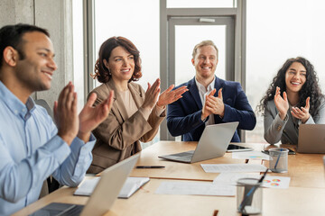 Poster - A group of diverse colleagues is sitting around a table in a modern office space. They are clapping and smiling, clearly pleased with the outcome of a successful meeting