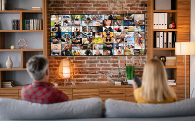 Canvas Print - A couple sits on a couch in a living room, facing a large television screen displaying a mosaic of images. The room features a brick wall and wood accents, suggesting a comfortable and cozy atmosphere