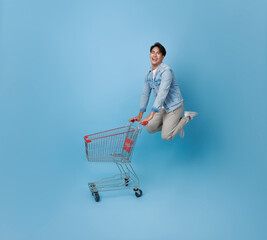 Full length happy excited Asian teen man pushing empty shopping cart or shopping trolley jumping promotion supermarket isolated on blue background.