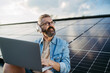 © Halfpoint - Man sitting on roof full of solar panels, headphones on head, typing on laptop.