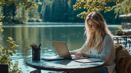 Poster - A woman is sitting at a table by a lake, working on her laptop. Concept of relaxation and tranquility, as the woman enjoys the peaceful surroundings while she works