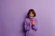 © Rattanapon - Satisfied smiling woman reads great message in her smartphone isolated on purple pastel background.