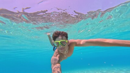 Wall Mural - Young man holding camera underwater swimming in turquoise water looking camera smiling having fun. Happy cheerful male enjoying summer
